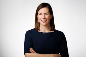 Melissa Moritz of STEM Next, a woman with straight brown hair, wearing a dark blue dress and a necklace, stands against a plain white background, smiling with her arms crossed.