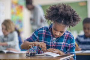 A young girl of African descent is indoors in her elementary school classroom. She is sitting at her desk and doing a science experiment with wires and circuits.