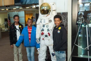 Three boys smile as they stand next to a white astronaut suit display in a museum setting, with glass cases and exhibits visible in the background.