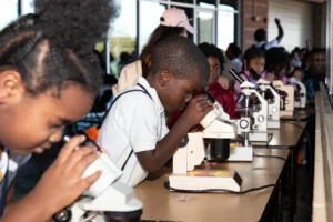 A group of children sits at a long table, each looking through a microscope—focused and engaged as they explore scientific samples, embodying youth STEM success stories in a brightly lit classroom or lab setting.