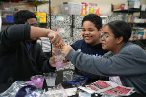 Three children wearing safety goggles smile and work together on a science experiment, mixing materials in a plastic bag at a classroom table with science supplies around them.
