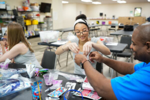 A smiling young woman and a man work together on a hands-on project at a table in a classroom during a casual STEM career exploration. Supplies, snacks, and craft materials are spread out, while others are seated and working in the background.