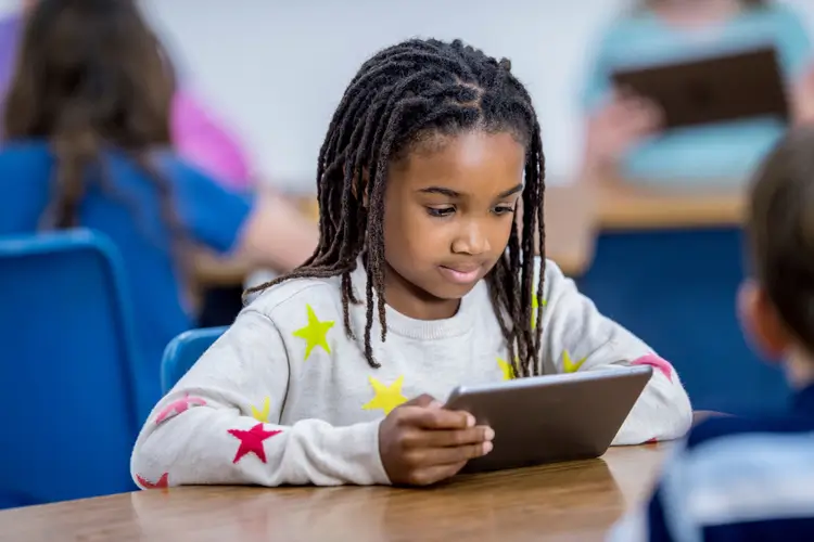 A young girl with long braided hair and a gray sweater with colorful stars sits at a desk in a classroom, focused on using a tablet device. Other children are visible in the background, also using tablets.