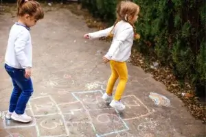 Two young girls play hopscotch on a sidewalk decorated with colorful chalk drawings. One girl jumps while the other waits her turn. Green bushes line the right side of the scene.