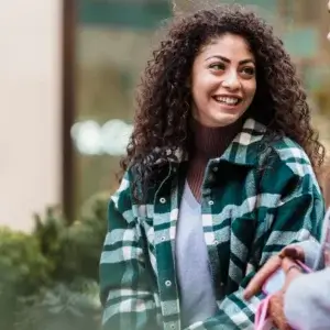 A woman with curly hair wearing a green and white plaid jacket smiles while talking to someone outdoors.