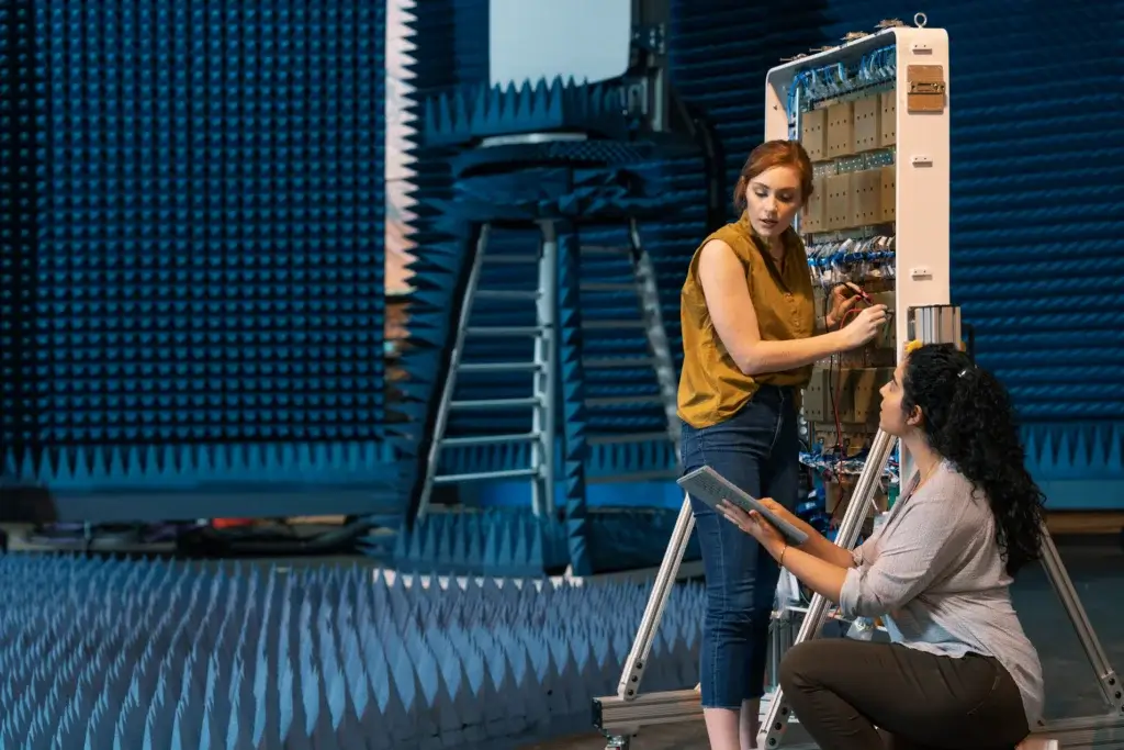 Two women work together on an electronic device in an anechoic chamber. One stands and adjusts wiring while the other kneels, holding a digital tablet. Blue sound-absorbing foam panels cover the room.