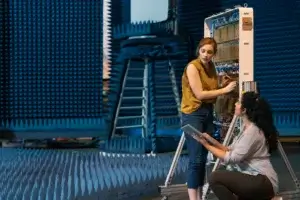 Two women work together on an electronic device in an anechoic chamber. One stands and adjusts wiring while the other kneels, holding a digital tablet. Blue sound-absorbing foam panels cover the room.