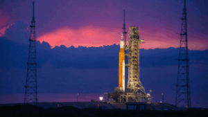 A rocket stands upright on a launch pad at dusk, illuminated by lights, with a dramatic purple and pink sunset sky and dark clouds in the background. Metal towers surround the launch structure.