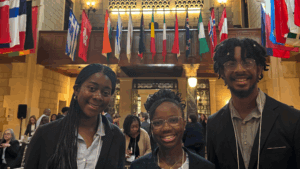 Three young adults in business attire smile at the camera in a large hall with international flags above, highlighting a youth development or STEM conference. Other attendees are gathered and seated in the background, suggesting a formal event.