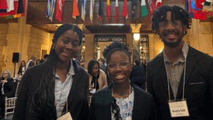 Three young adults dressed in business attire, wearing name badges, smile for a photo in a conference room filled with people. International flags hang from the ceiling, highlighting a global event focused on youth development and STEM.