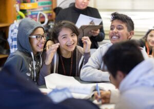 A group of smiling students sit together at a table in a classroom during an afterschool summer STEM session, laughing and talking while doing schoolwork. Books and papers are spread in front of them, as a teacher stands in the background holding a book.