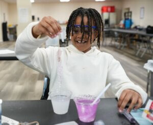 A smiling child wearing safety goggles and a white hoodie stretches a strand of slime between two cups, enjoying an afterschool summer STEM activity at a table in a classroom with science materials around.