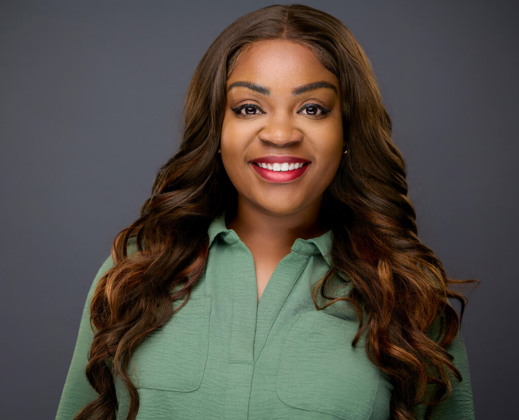 A woman with long wavy brown hair and a green collared shirt smiles in front of a plain dark gray background.