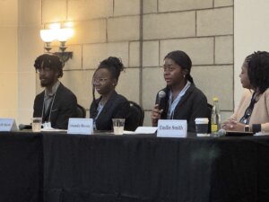 Four people sit at a panel table, three women and one man, all dressed in business attire. One woman speaks into a microphone. Name cards, drinks, and papers are on the table in front of them.