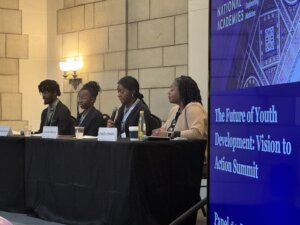 Four panelists speak at a table during a summit. A large screen beside them displays the event title: “The Future of Youth Development: Vision to Action Summit.” Microphones and drinks are on the table.