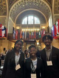 Three people wearing business attire and name tags smile at the camera inside a grand hall with arched windows and many international flags hanging from the ceiling. Other attendees and ornate details are visible in the background.