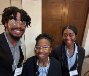 Three people wearing business attire and name tags stand indoors, smiling for the camera. In the background, a wooden door and light-colored stone wall hint at a professional setting focused on youth development in STEM.