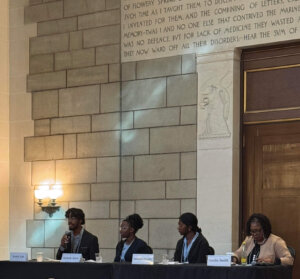 Four panelists sit at a table on a stage, speaking to an audience in a formal room with stone walls and a tall inscription. People are seated in gold chairs, listening attentively to the discussion.