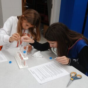 Two girls conduct a science experiment, carefully pouring liquid into test tubes on a white table. A sheet of instructions and scissors are nearby. Both are focused on their task.