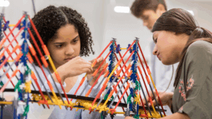 Two students focus on building a bridge structure with colorful plastic connecting rods and gears in a classroom, while another student works in the background.