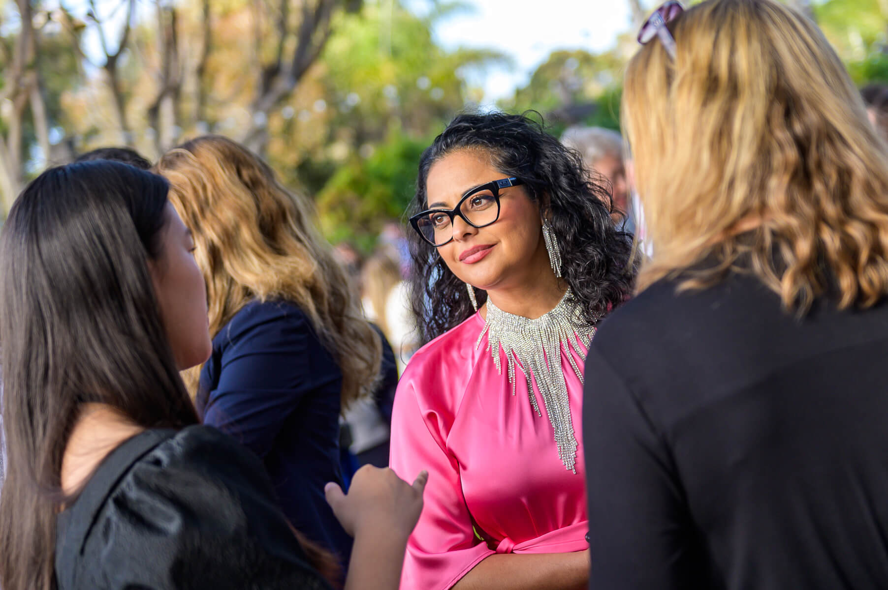 A woman in a pink dress and glasses listens attentively while talking to two other women at an outdoor event with trees in the background.
