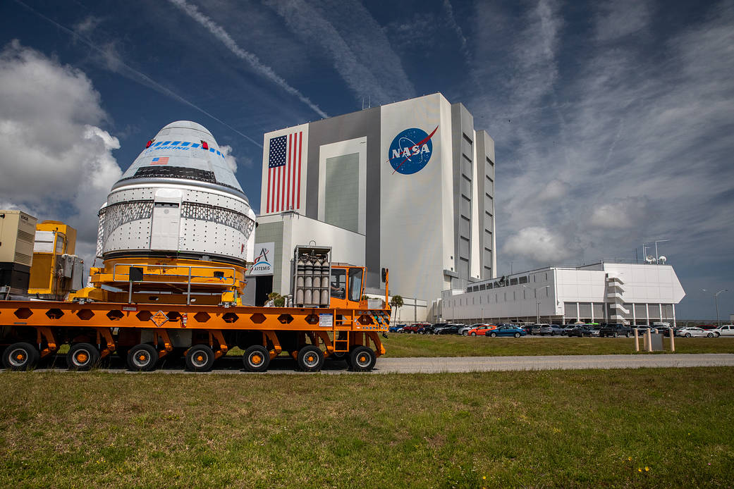 A spacecraft on a yellow transport vehicle is moved outside NASA’s Kennedy Space Center, with the Vehicle Assembly Building and a large NASA logo visible in the background under a partly cloudy sky.