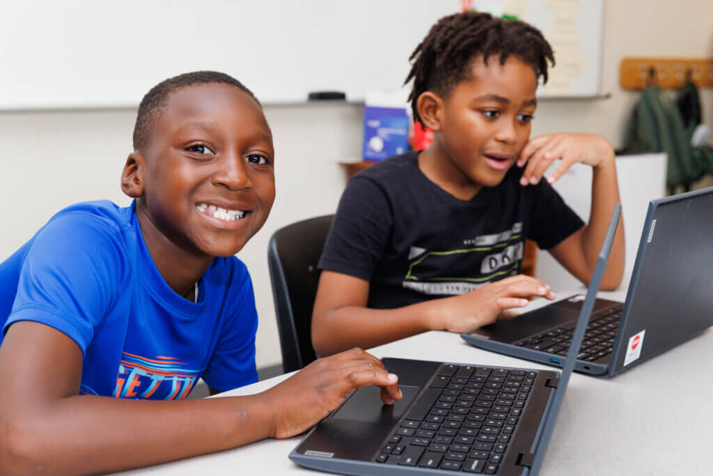 Two young boys sit at a table in a classroom, each using a laptop. The boy in the blue shirt smiles at the camera while the other, focused on his screen, explores AI learning in afterschool activities.