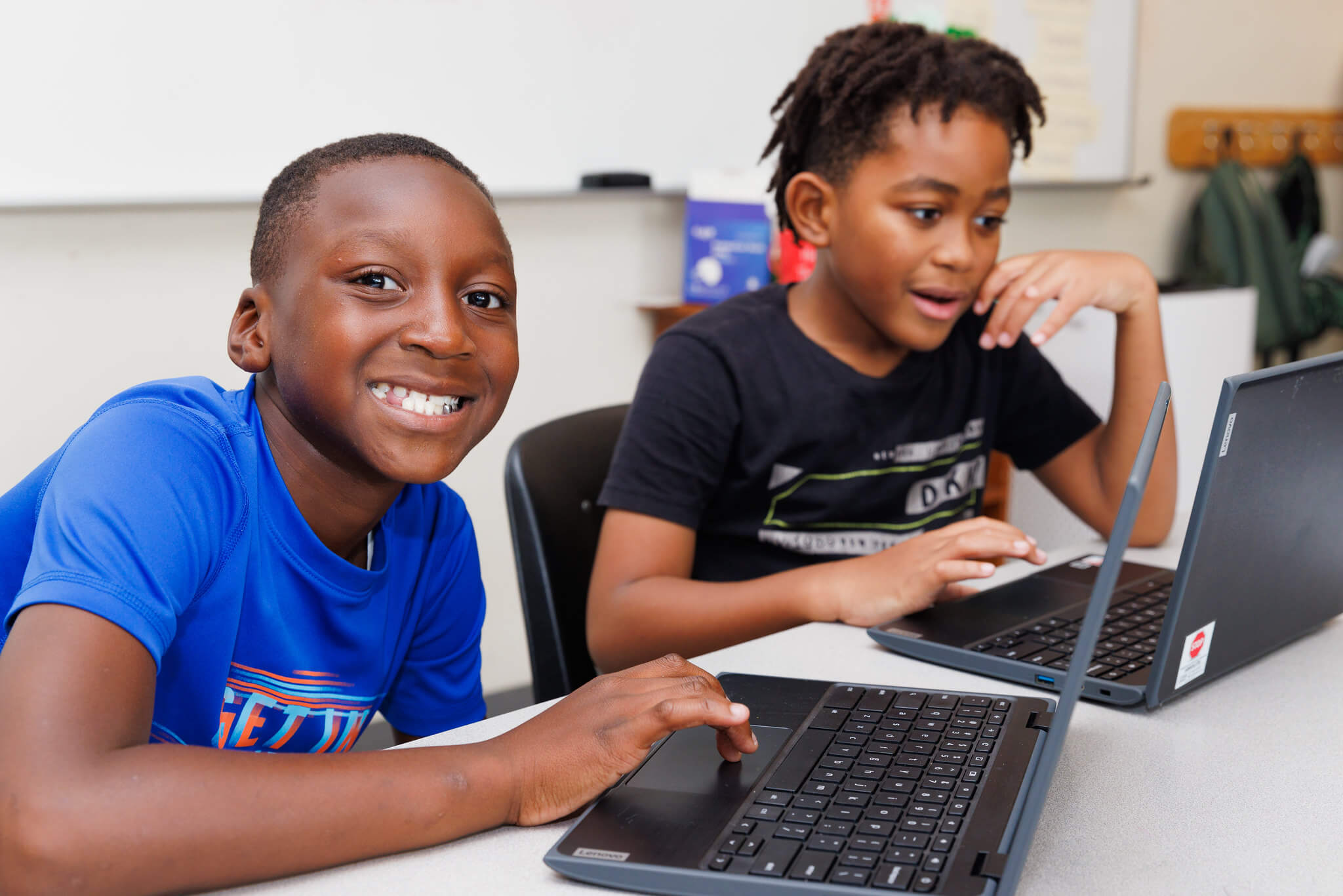 Two young boys sit at a table in a classroom, each using a laptop. The boy in the blue shirt smiles at the camera while the other, focused on his screen, explores AI learning in afterschool activities.