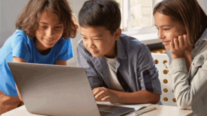 Three children sit together at a table, smiling and looking at a laptop screen. Sunlight comes through a window behind them, and a notebook with a pen lies on the table nearby.