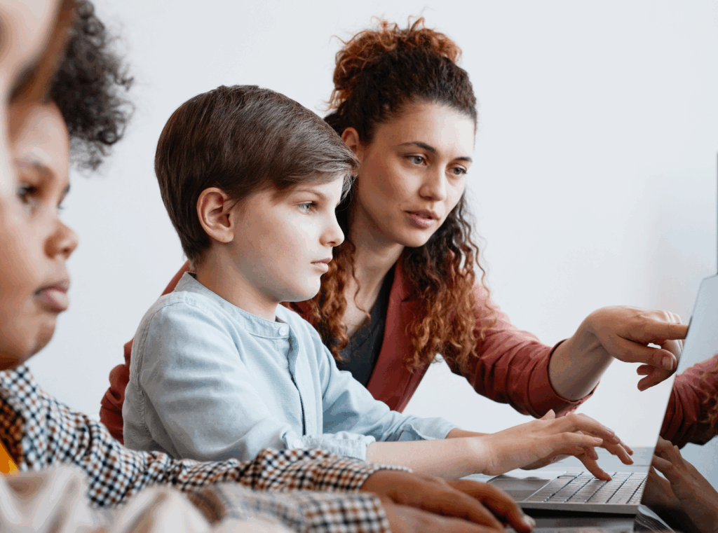 A teacher helps a young boy using a laptop in a classroom during afterschool STEM learning. Other children are nearby, also using laptops. A screen in the background displays the word Digital.