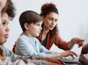 A teacher helps a young boy using a laptop in a classroom during afterschool STEM learning. Other children are nearby, also using laptops. A screen in the background displays the word Digital.