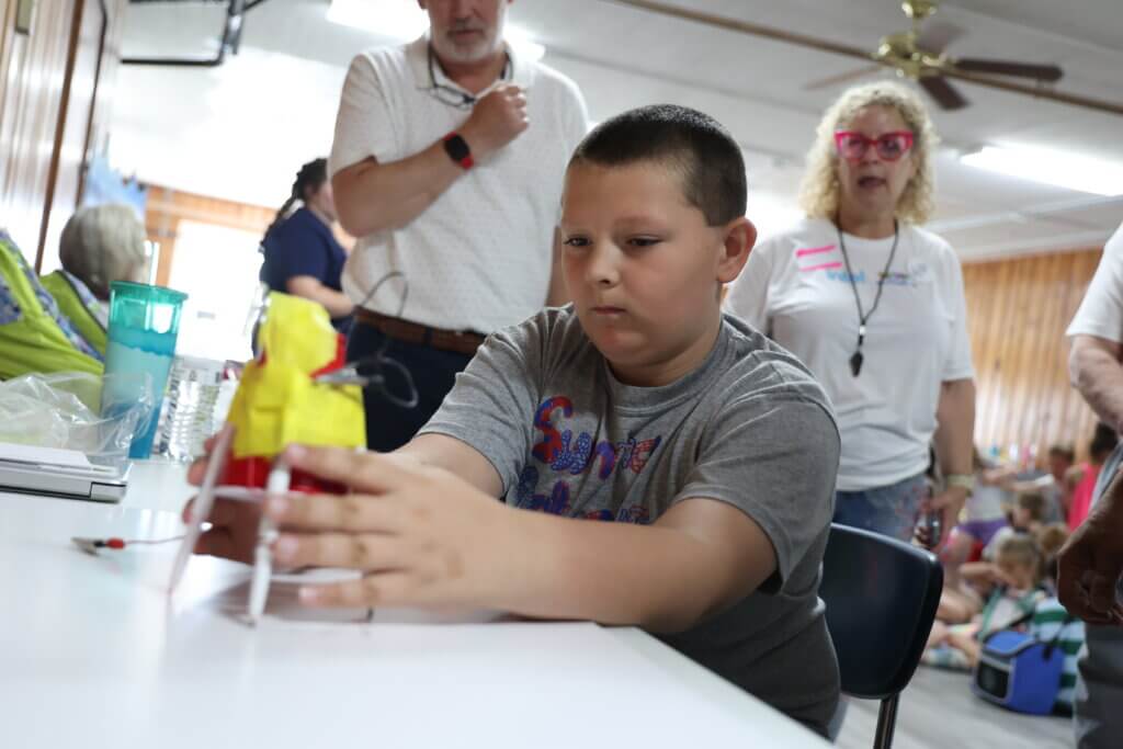 A young boy sits at a table working with a small robot model, engaging in STEM activities while adults and children watch in the background inside a room with wood-paneled walls.