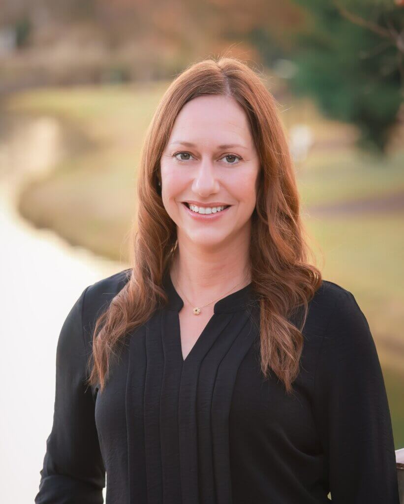 A woman with long, light brown hair and a black blouse smiles outdoors by a river or lake, with trees and blurred greenery in the background.
