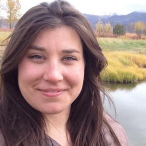 A woman with long brown hair smiles outdoors near a calm body of water, with grass, trees, and distant mountains in the background. The sky appears partly cloudy.