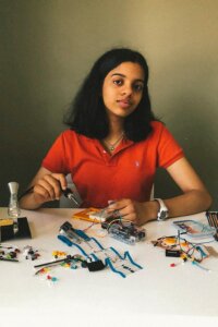A young woman in an orange shirt sits at a table with electronic components, wires, and a soldering iron, appearing to work on a circuit project that promotes AI literacy.