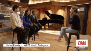 Four young people sit on stools in a recording studio, discussing AI literacy with a host. A grand piano is in the background. The screen reads: Neha Shukla | WAFF Youth Delegate and CNN CONNECT THE WORLD.