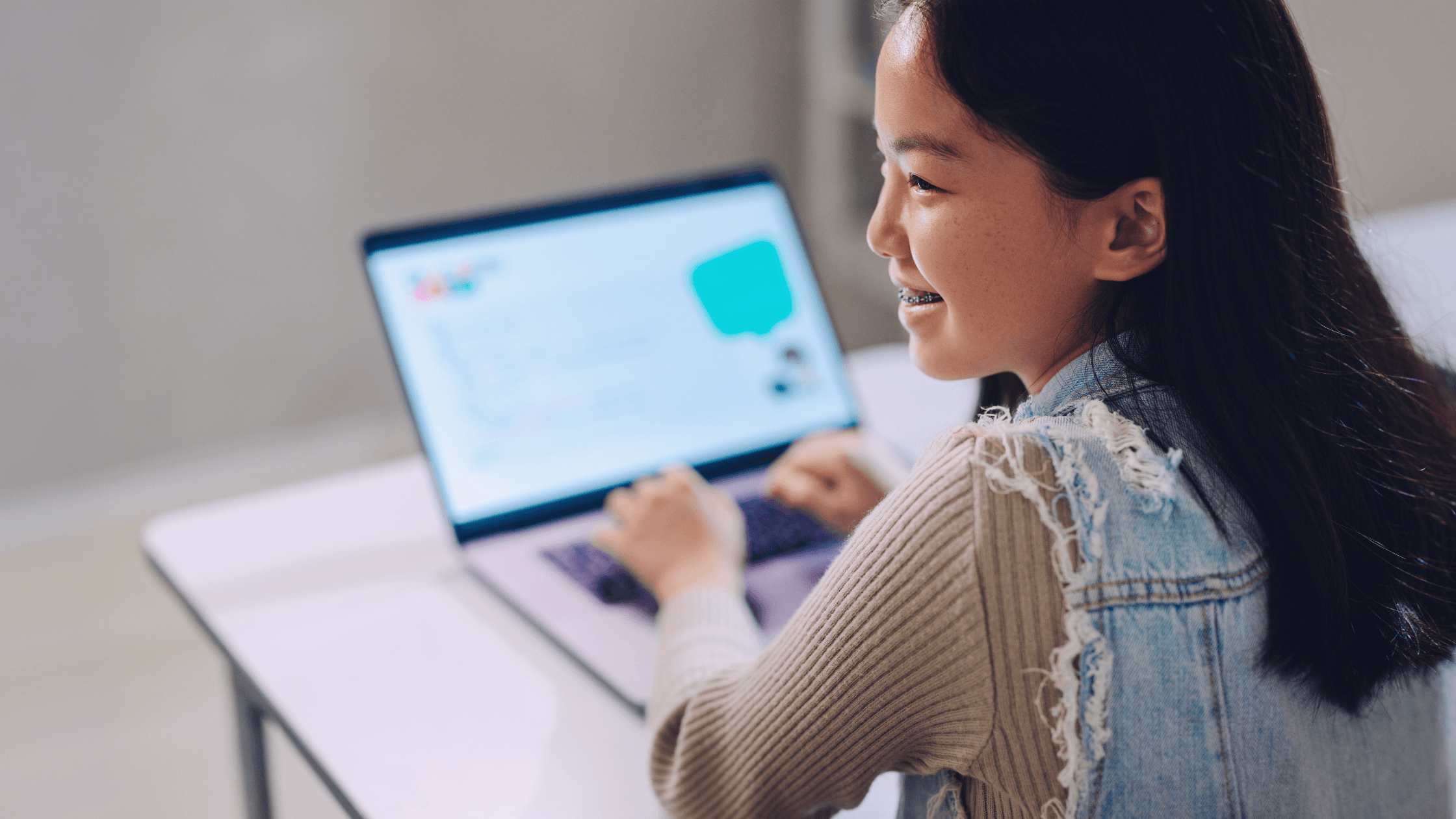 A smiling girl sits at a desk using a laptop, wearing a light denim vest over a sweater. The computer screen shows a chat or coding interface. She appears to be engaged and focused.