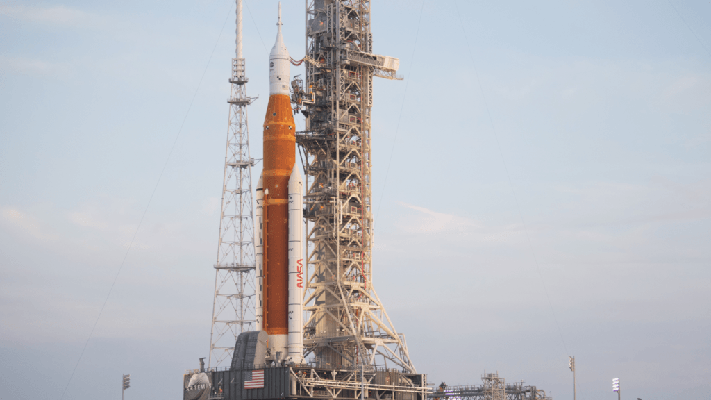 A large orange and white NASA rocket stands upright on a launch pad, next to a tall metal launch tower, against a light blue sky with some clouds.