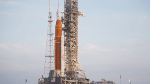 A large orange and white NASA rocket stands upright on a launch pad, next to a tall metal launch tower, against a light blue sky with some clouds.