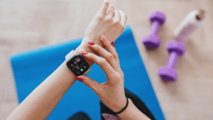 A person with pink nail polish checks a smartwatch showing fitness stats while sitting on a blue exercise mat, with purple dumbbells and a water bottle nearby.