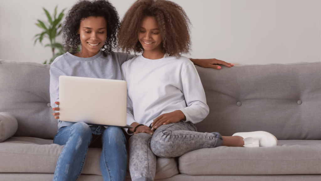 Two women with curly hair sit close together on a gray couch, smiling and looking at a laptop. One has her arm around the other. There is a green plant in the background.