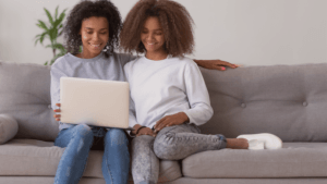 Two women with curly hair sit close together on a gray couch, smiling and looking at a laptop. One has her arm around the other. There is a green plant in the background.
