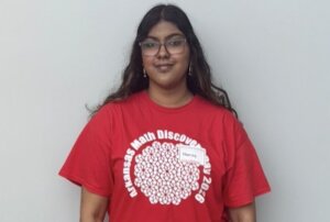 A person with long dark hair and glasses stands in front of a gray wall, wearing a red Arkansas Math Discovery Day 2024 T-shirt and a name tag that reads Hansa.