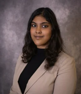 A young woman with wavy dark hair in a beige blazer and black top poses for a professional portrait against a gray, textured background.