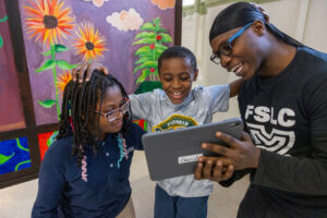 A man and two smiling children look at a tablet together in a colorful room with a flower mural. The man gently rests his hand on one child’s head as they all enjoy what’s on the screen.