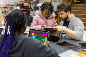 A man helps a young girl in a pink coat use a tablet, while another child with purple braids works on a colorful music app on a tablet at a classroom table.