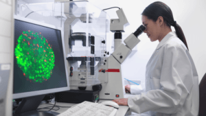 A scientist in a white lab coat looks into a microscope in a laboratory. A computer screen nearby displays a colorful, circular microscopic image with green and red spots.