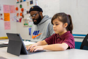 A young girl in a maroon shirt types on a tablet at a desk, while an adult man wearing glasses and a beanie sits next to her, looking at papers in a classroom setting.