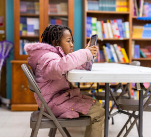 A young girl in a pink coat sits at a round table in a library or classroom, focused on her tablet as she explores AI literacy, with bookshelves full of books in the background.