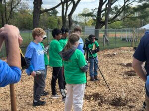 A group of kids in green shirts stands outdoors on wood chips, watching a peer operate a video camera on a tripod. Trees, fencing, and more people are visible in the background.